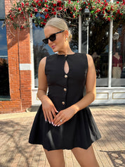 Woman wearing fit to flare black mini dress with keyhole cutout and gold buttons.