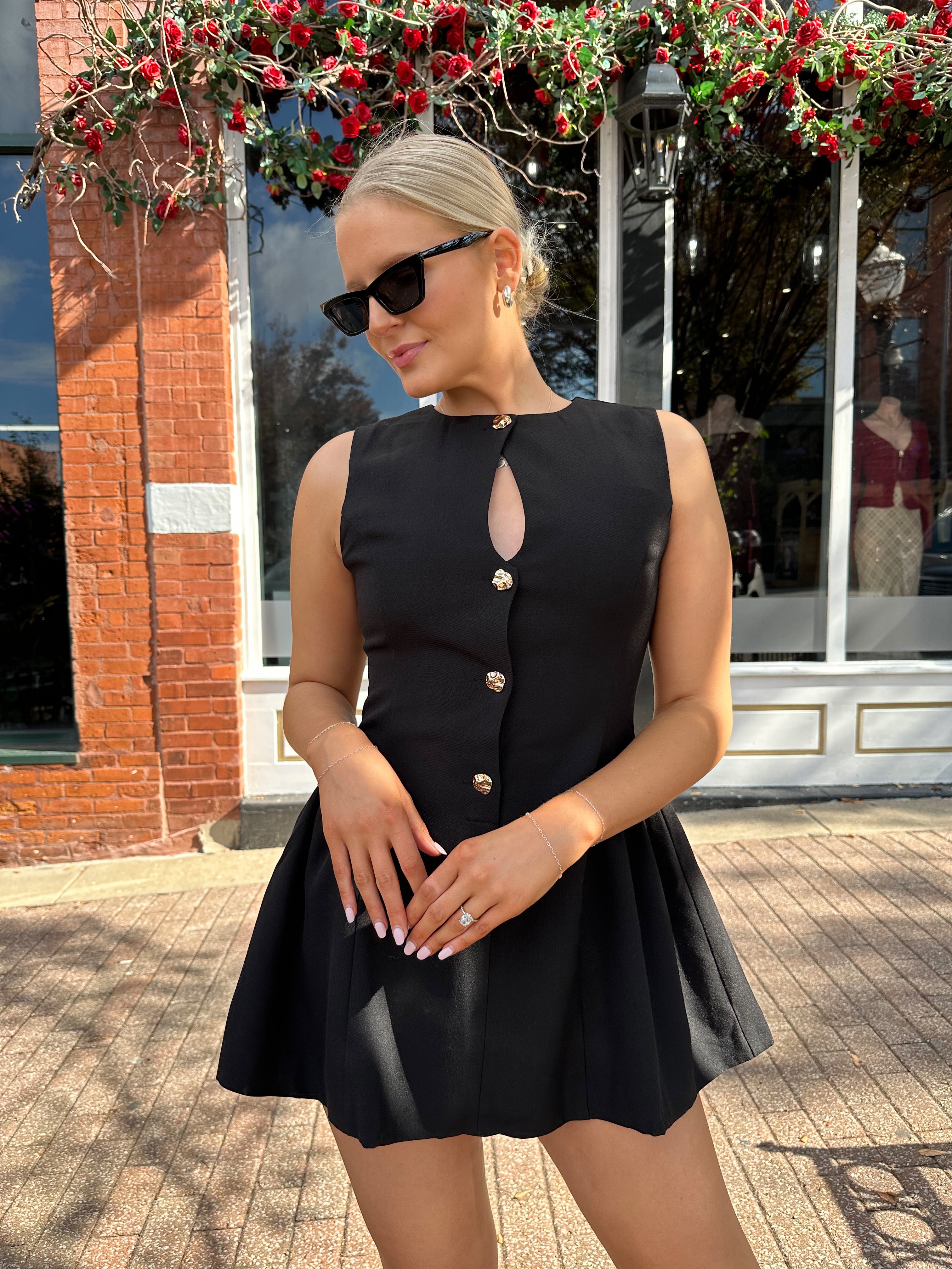 Woman wearing fit to flare black mini dress with keyhole cutout and gold buttons.