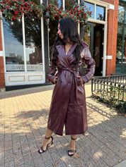 Woman wearing burgundy leather trench coat with belt and and button closure.
