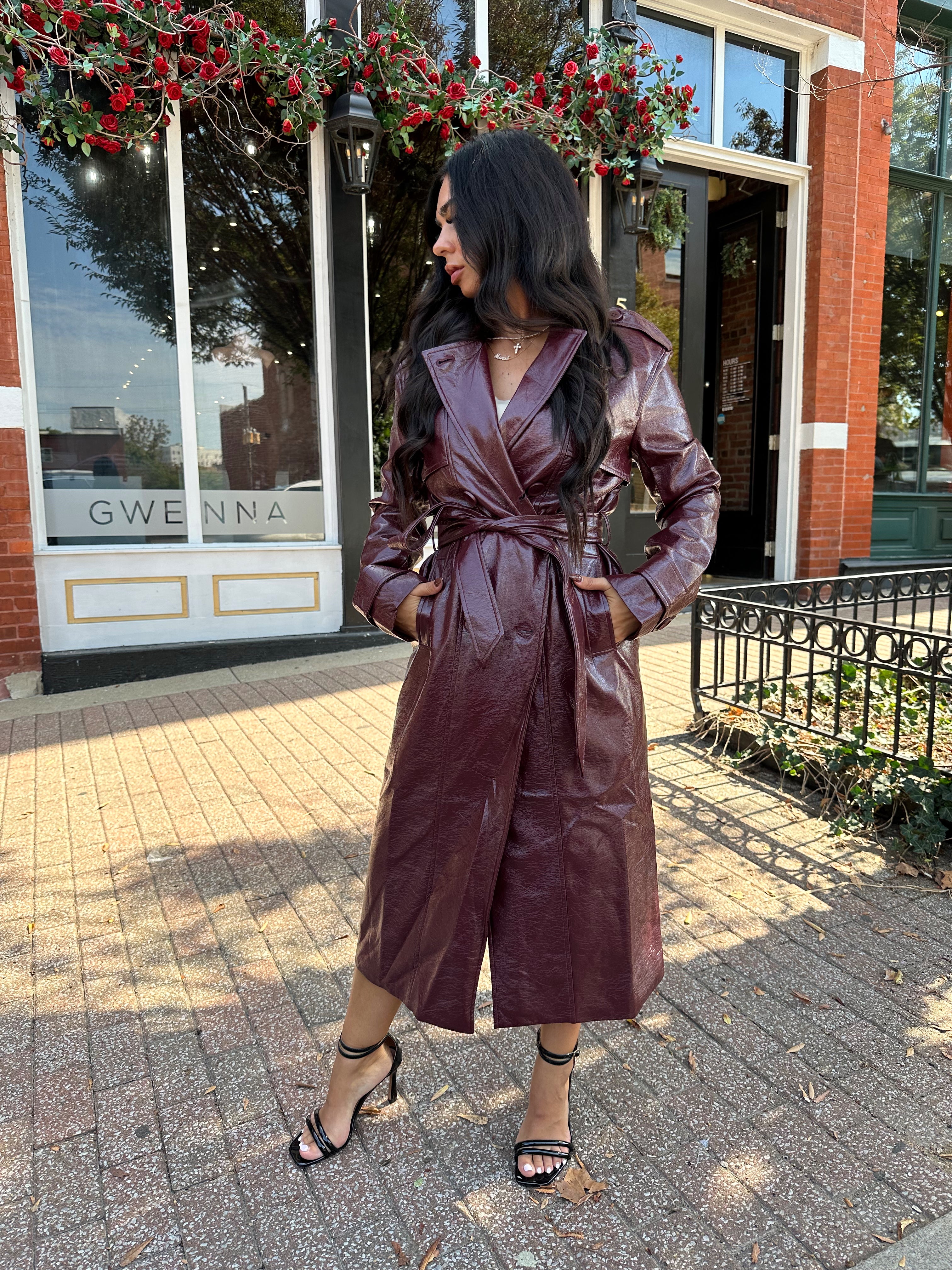Woman wearing burgundy leather trench coat with belt and and button closure.
