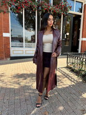 Woman wearing burgundy leather trench coat with belt and and button closure.