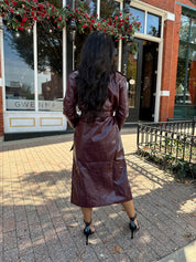 Woman wearing burgundy leather trench coat with belt and and button closure.