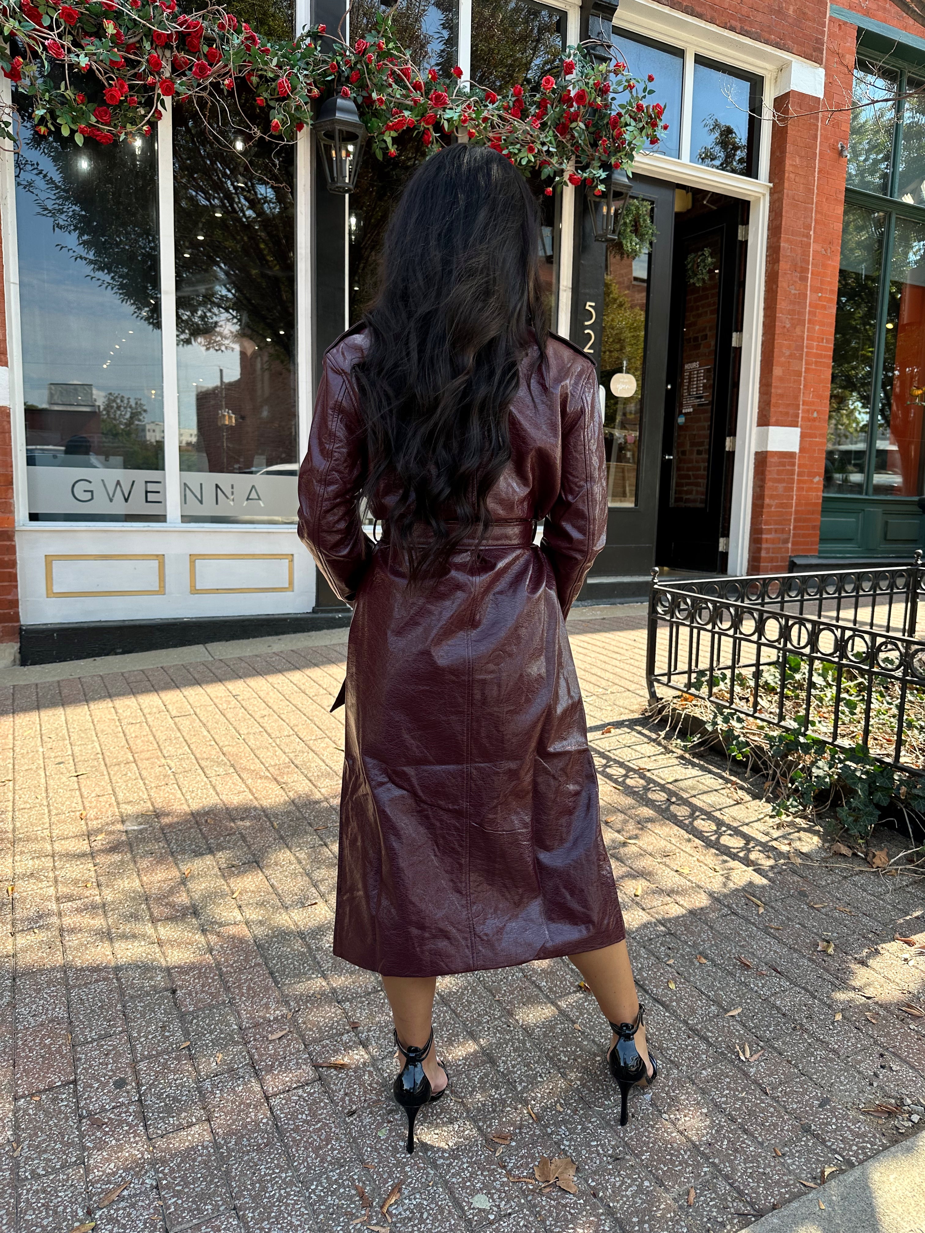 Woman wearing burgundy leather trench coat with belt and and button closure.