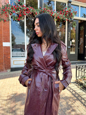Woman wearing burgundy leather trench coat with belt and and button closure.