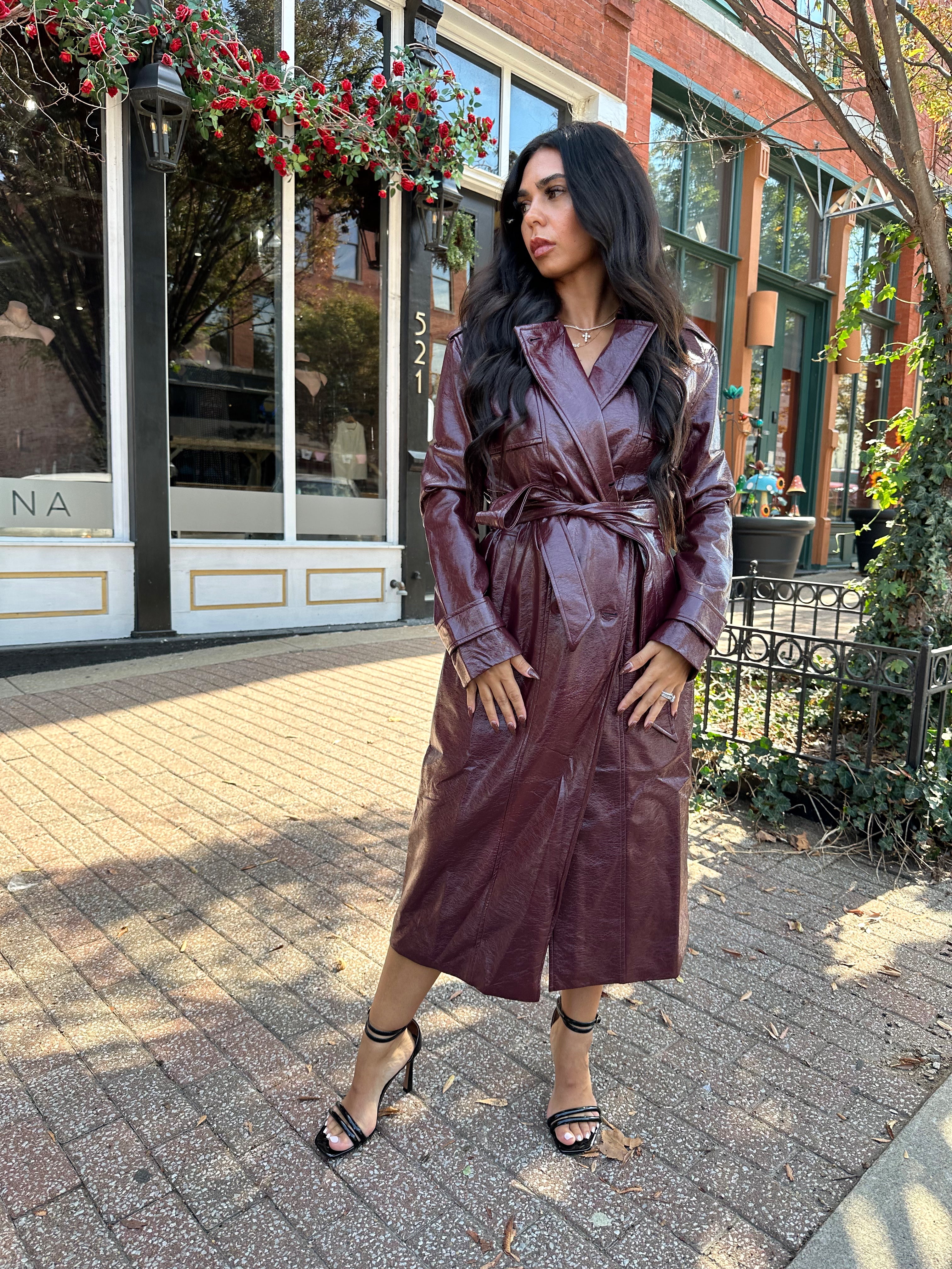 Woman wearing burgundy leather trench coat with belt and and button closure.