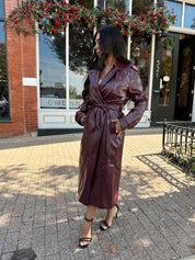 Woman wearing burgundy leather trench coat with belt and and button closure.