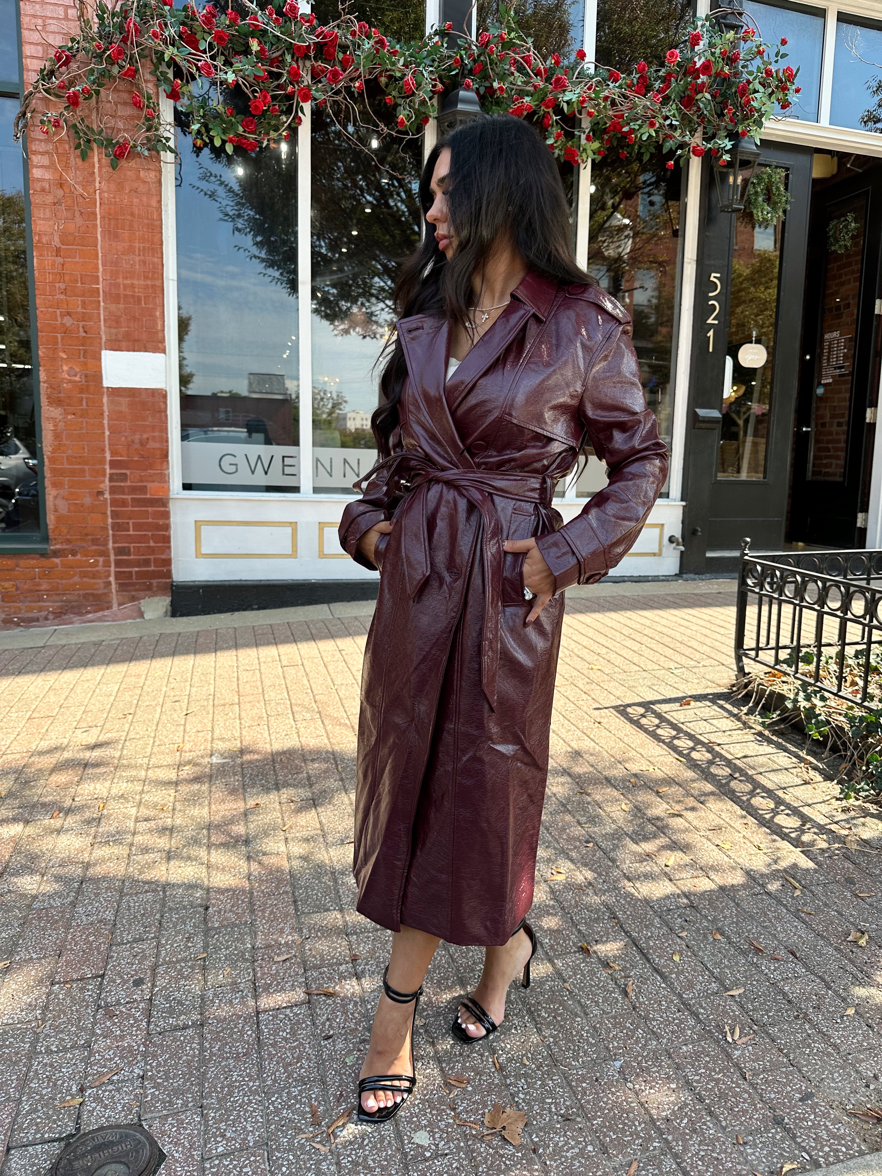 Woman wearing burgundy leather trench coat with belt and and button closure.