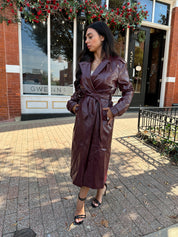 Woman wearing burgundy leather trench coat with belt and and button closure.