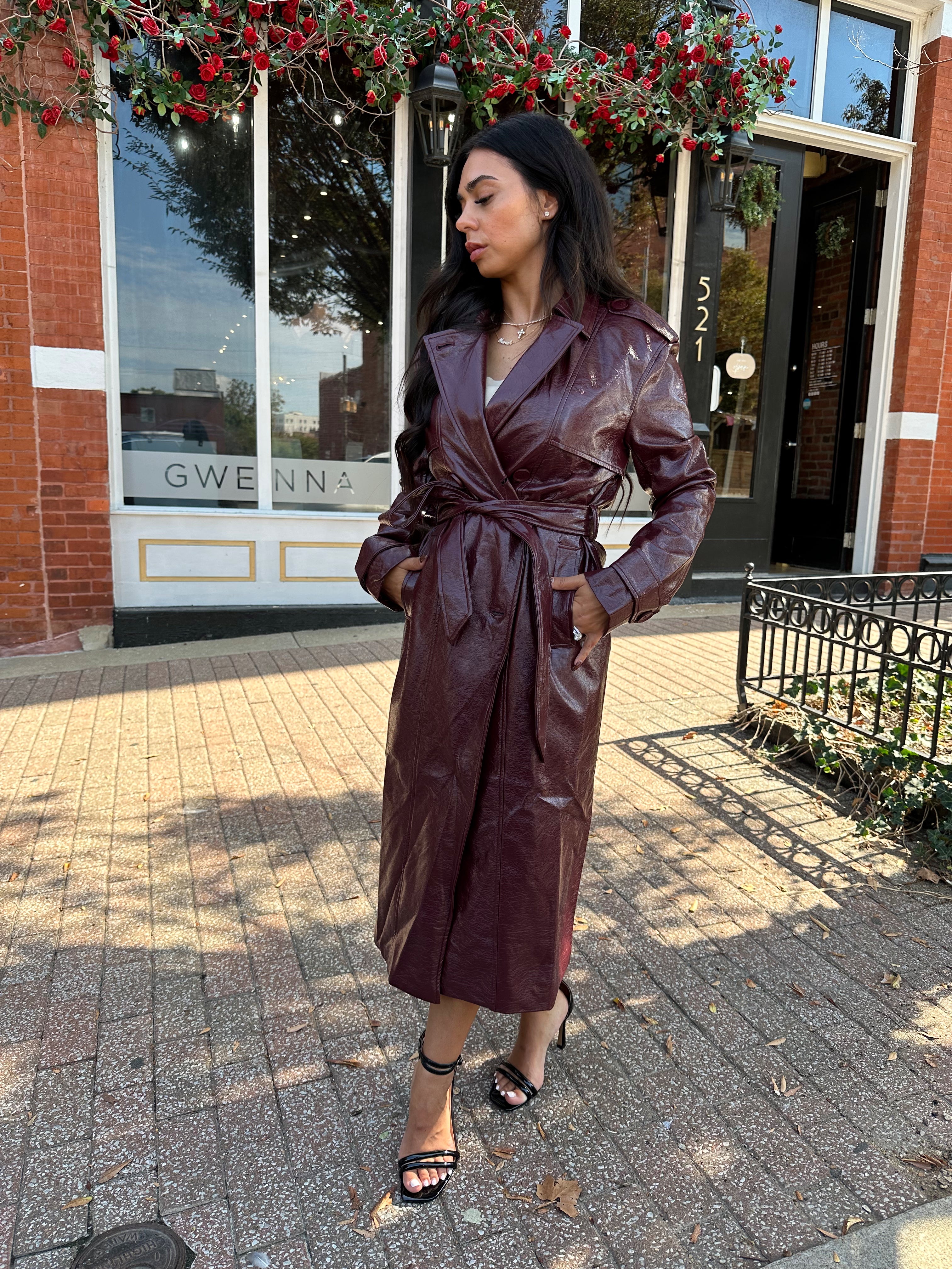 Woman wearing burgundy leather trench coat with belt and and button closure.