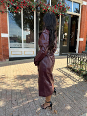 Woman wearing burgundy leather trench coat with belt and and button closure.