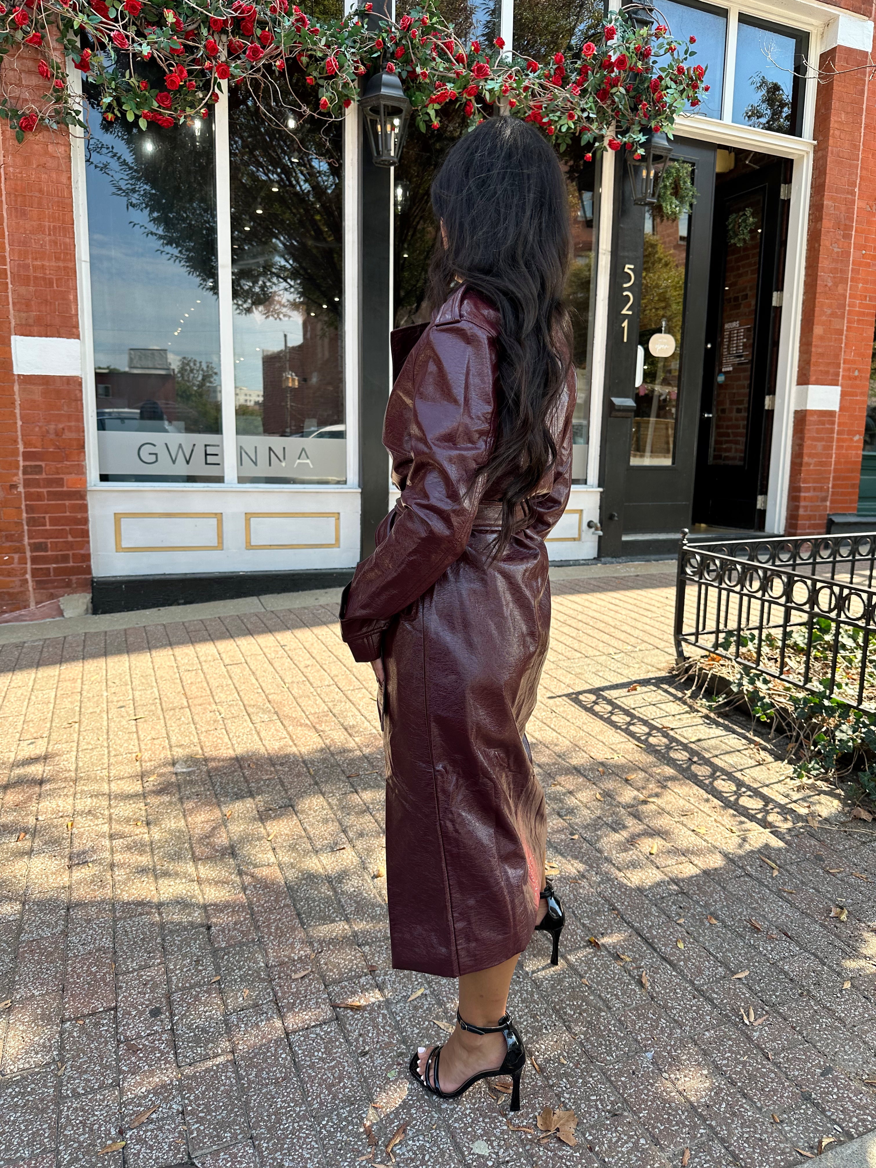 Woman wearing burgundy leather trench coat with belt and and button closure.