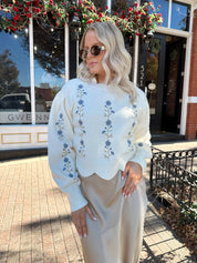 Woman wearing cream sweater with blue embroidered floral pattern.