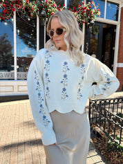 Woman wearing cream sweater with blue embroidered floral pattern.
