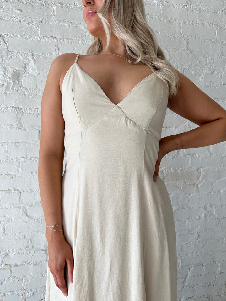 Woman wearing beige linen midi dress with an open tie back, and functional pockets.