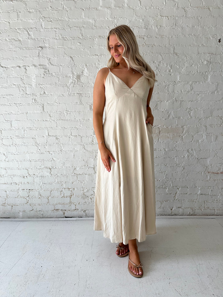 Woman wearing beige linen midi dress with an open tie back, and functional pockets.