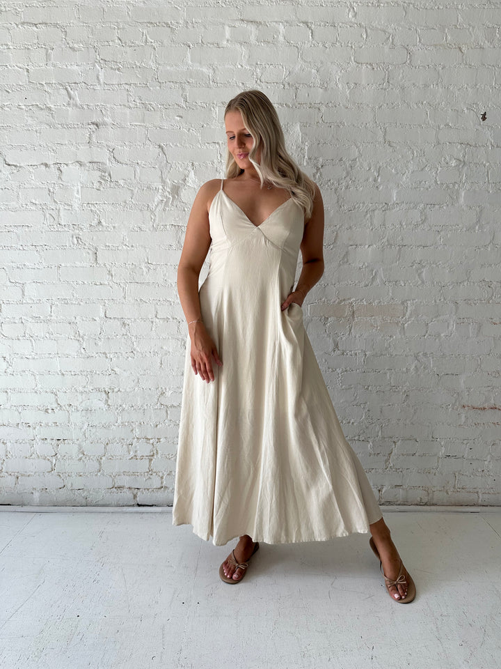Woman wearing beige linen midi dress with an open tie back, and functional pockets.