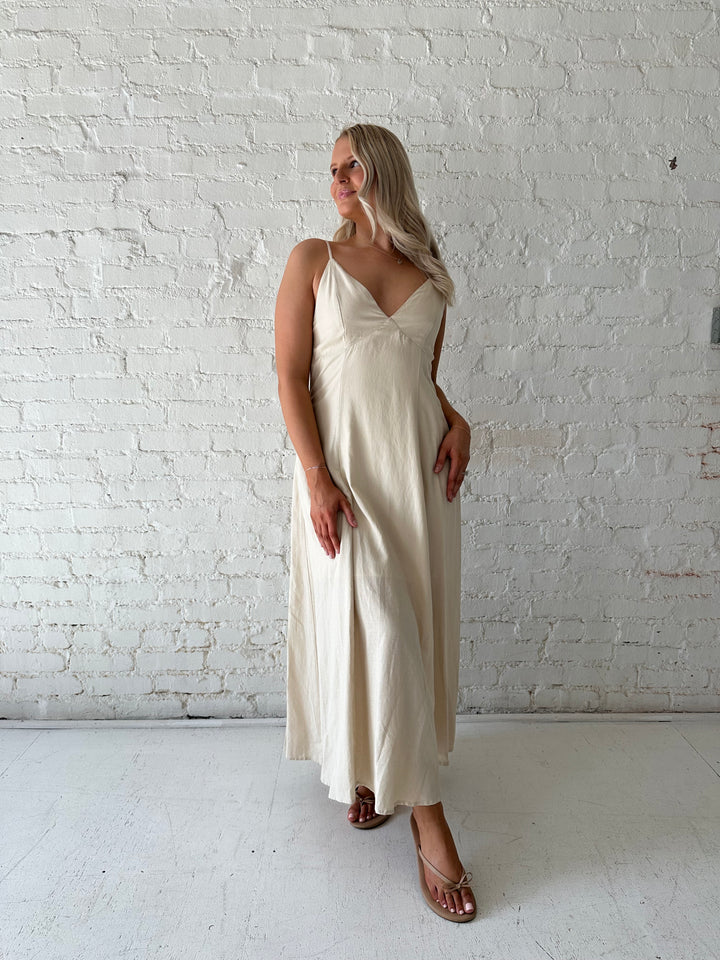 Woman wearing beige linen midi dress with an open tie back, and functional pockets.
