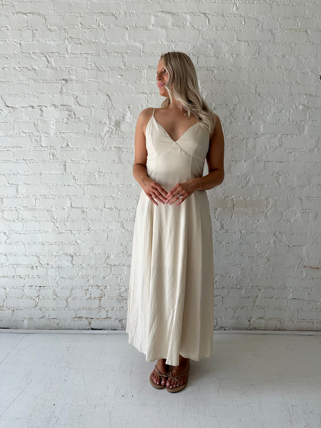 Woman wearing beige linen midi dress with an open tie back, and functional pockets.