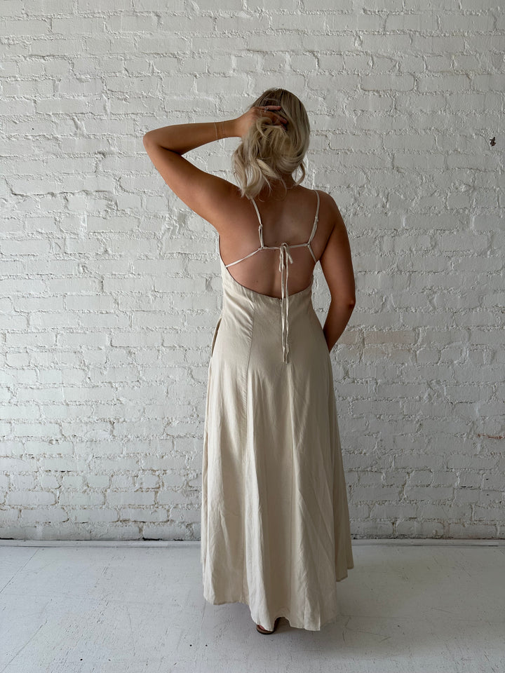 Woman wearing beige linen midi dress with an open tie back, and functional pockets.
