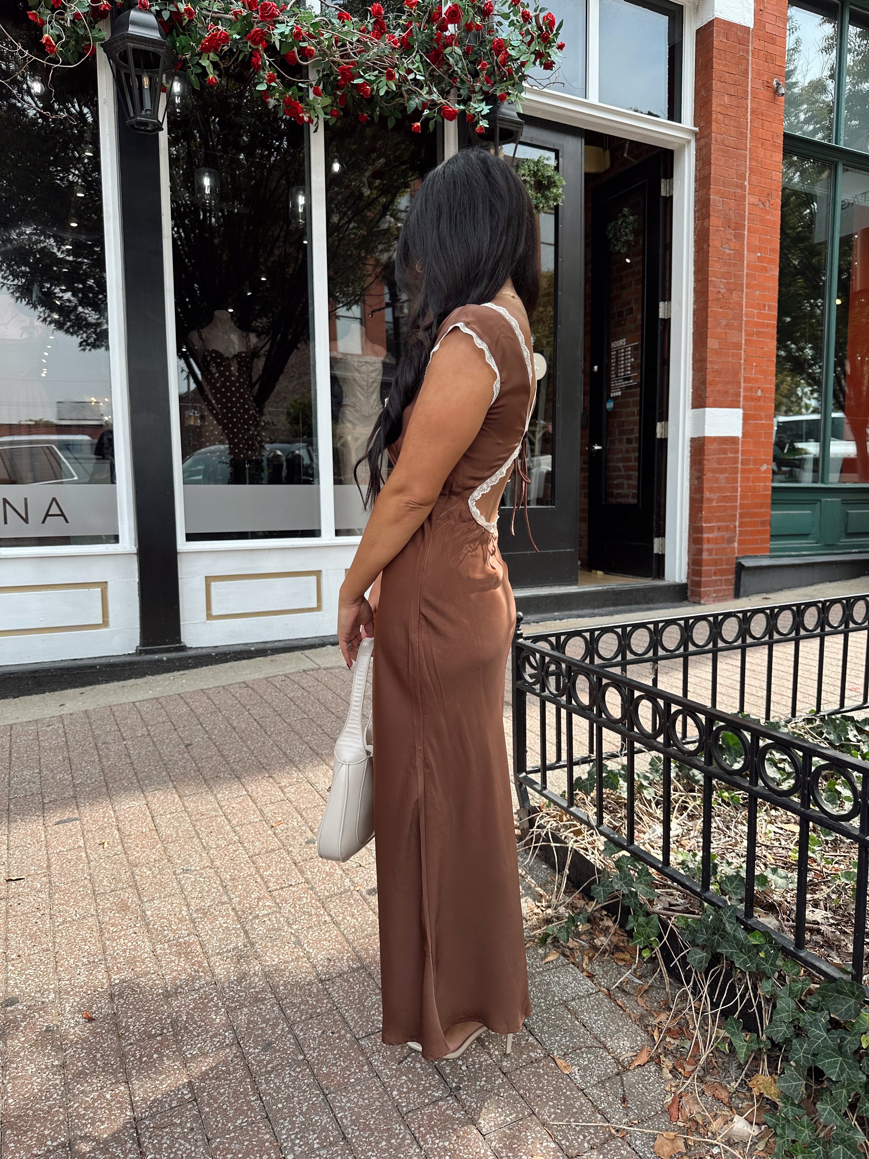Woman wearing copper brown satin midi dress with white ruffle trim and tie back. 
