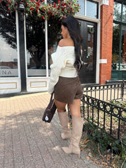 Woman wearing ivory long sleeve off the shoulder button up cardigan and brown suede shorts. 