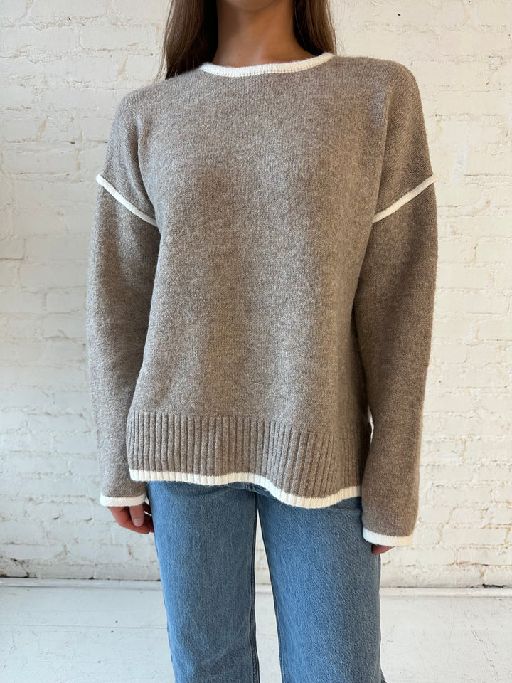 Woman wearing taupe knit sweater with contrast trim and medium wash jeans. 