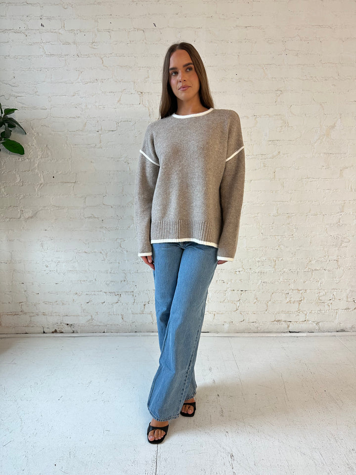 Woman wearing taupe knit sweater with contrast trim and medium wash jeans. 