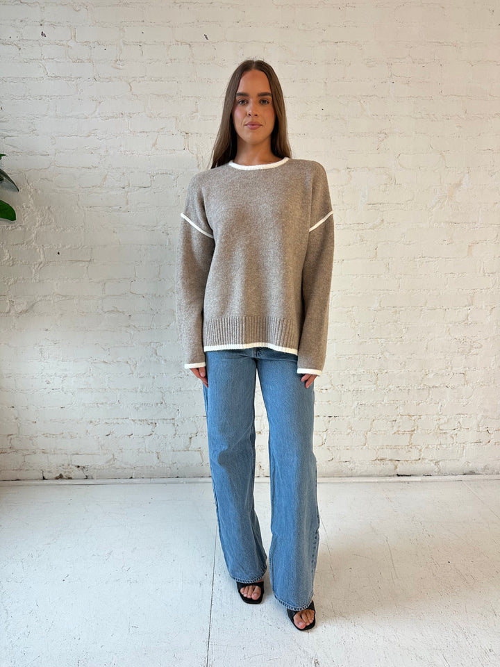 Woman wearing taupe knit sweater with contrast trim and medium wash jeans. 