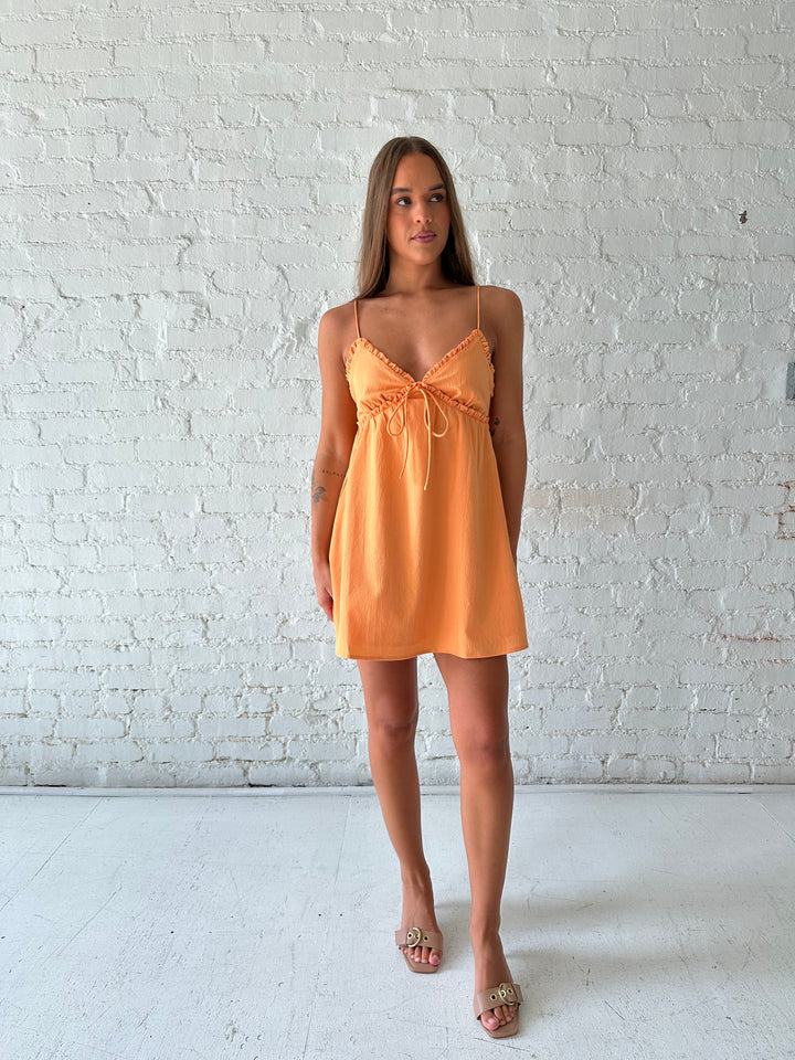 Woman wearing orange mini dress with ruffle trim neckline and tie detail. 