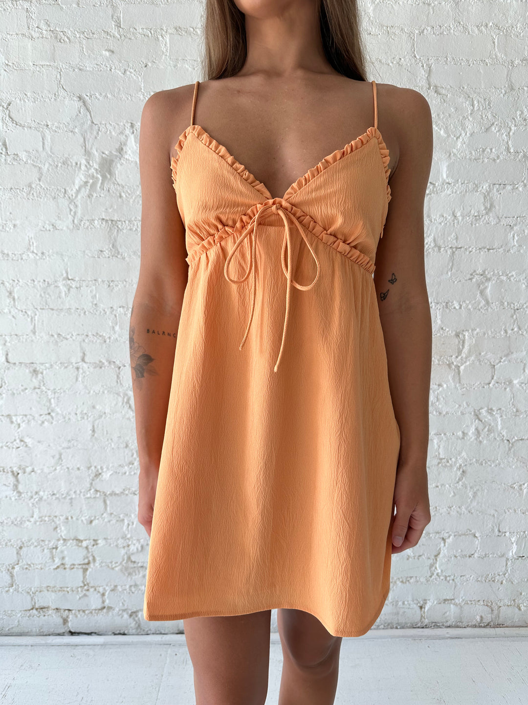 Woman wearing orange mini dress with ruffle trim neckline and tie detail.