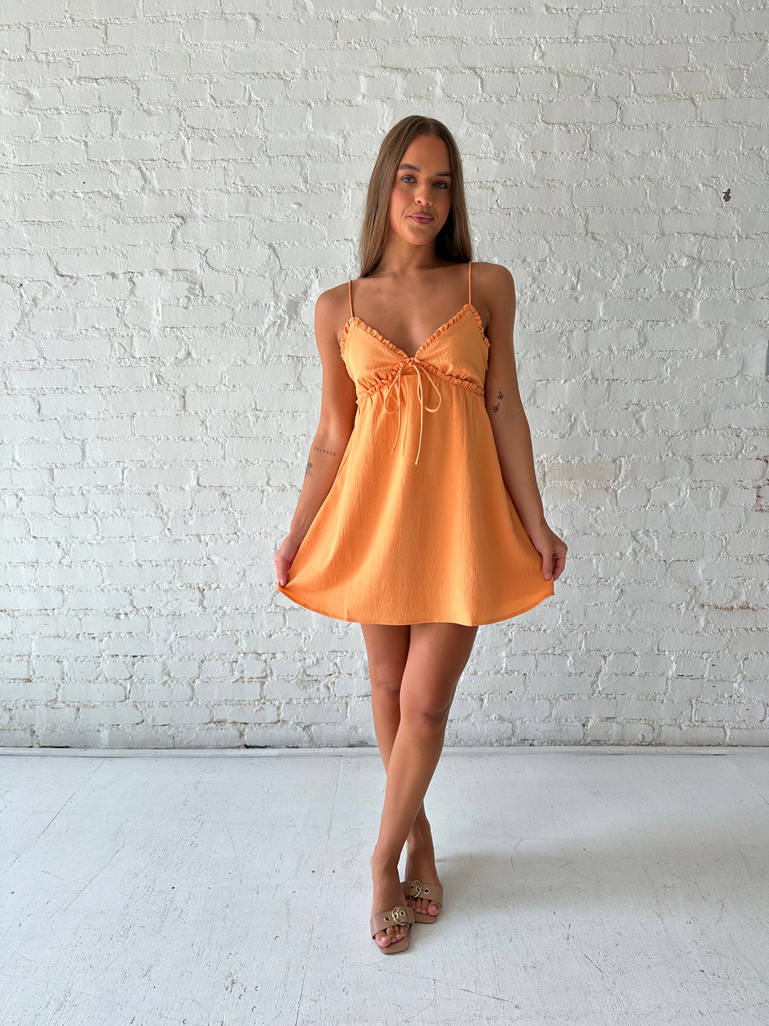 Woman wearing orange mini dress with ruffle trim neckline and tie detail.