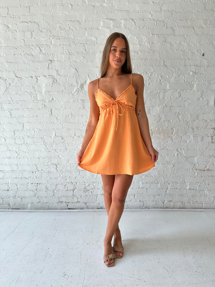 Woman wearing orange mini dress with ruffle trim neckline and tie detail.