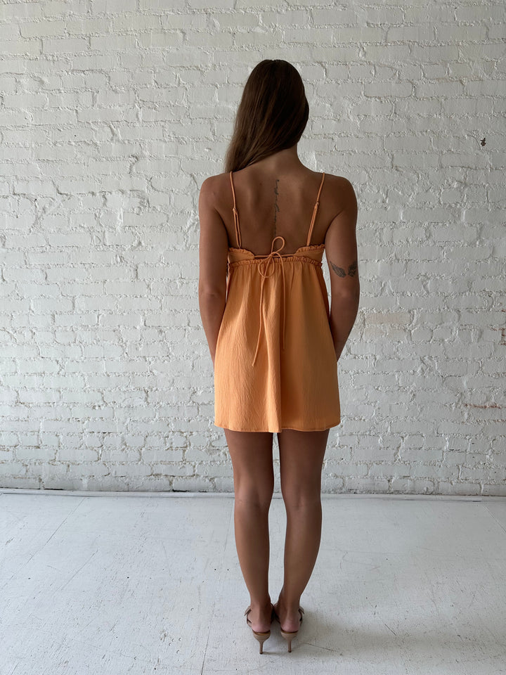 Woman wearing orange mini dress with ruffle trim neckline and tie detail.