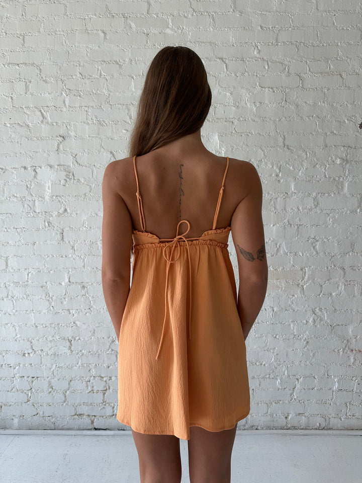 Woman wearing orange mini dress with ruffle trim neckline and tie detail.