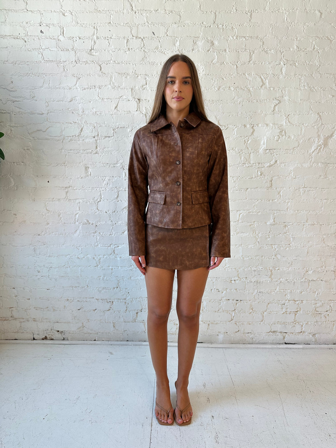 Woman wearing brown leather two piece set with tailored jacket top, and high waisted mini skort bottom.