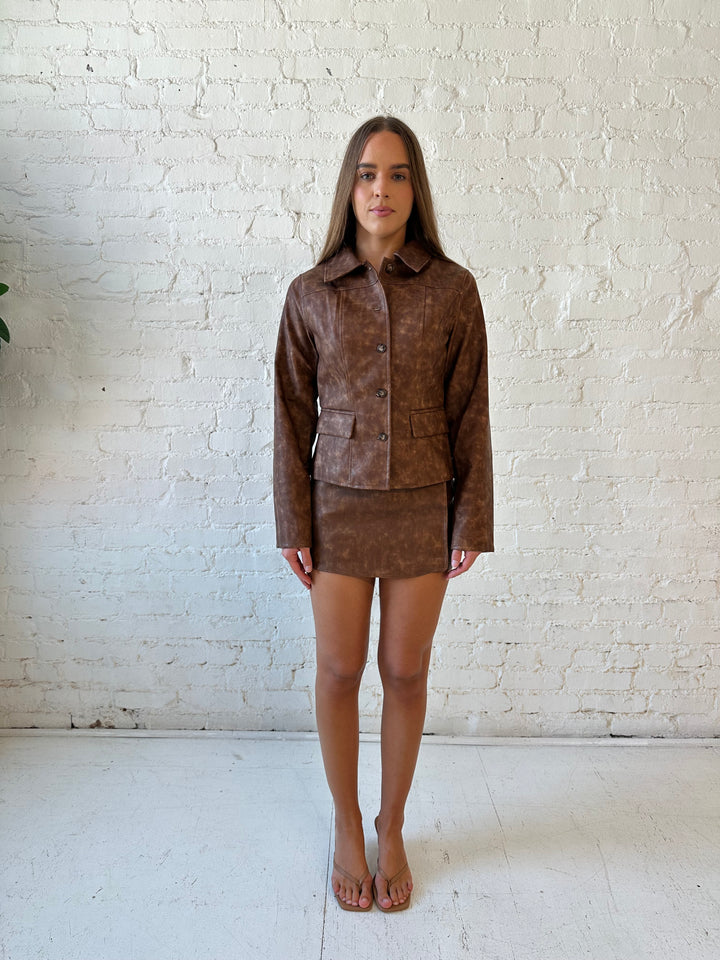 Woman wearing brown leather two piece set with tailored jacket top, and high waisted mini skort bottom.