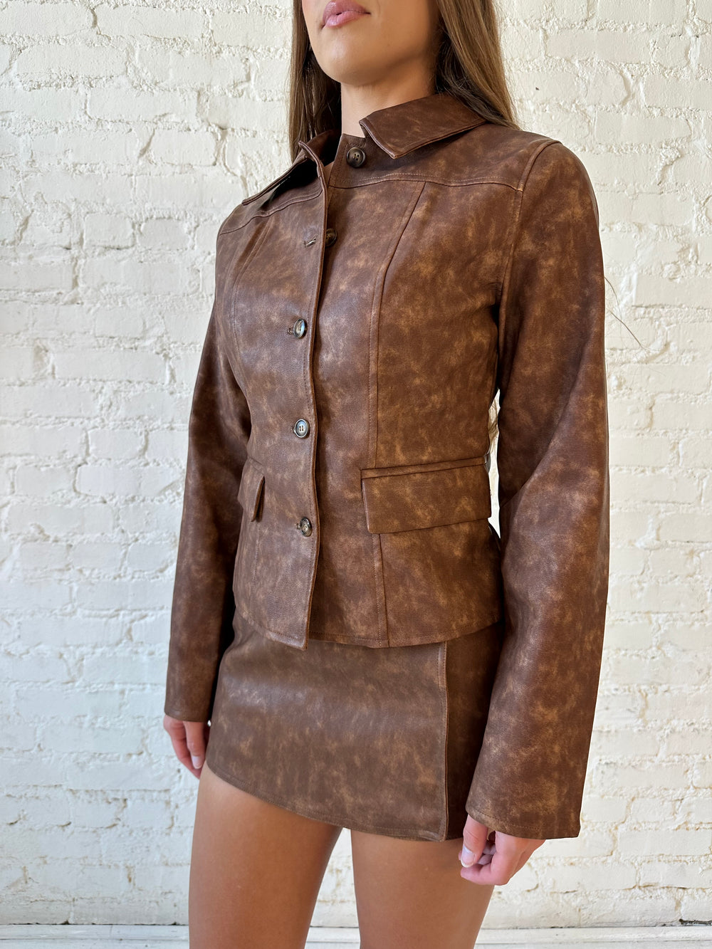 Woman wearing brown leather two piece set with tailored jacket top, and high waisted mini skort bottom.