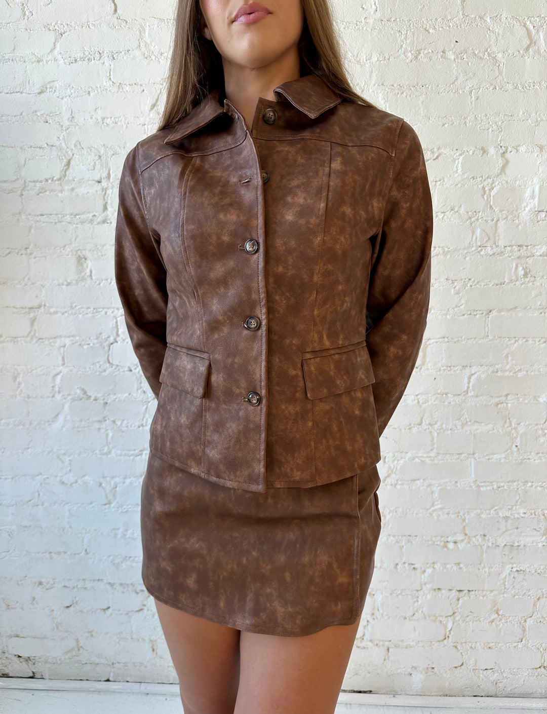 Woman wearing brown leather two piece set with tailored jacket top, and high waisted mini skort bottom.