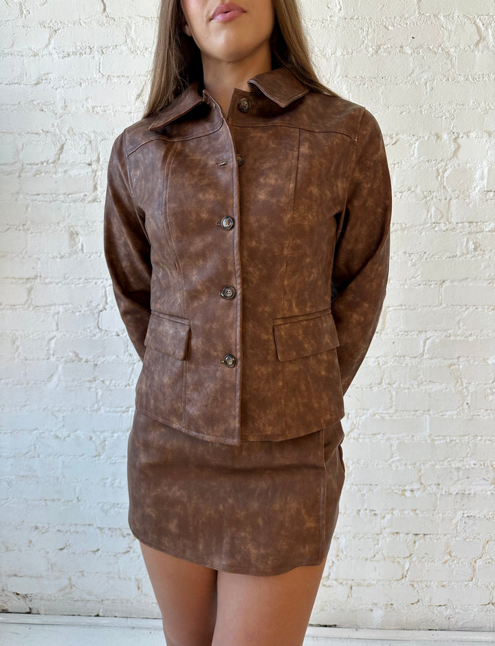 Woman wearing brown leather two piece set with tailored jacket top, and high waisted mini skort bottom.