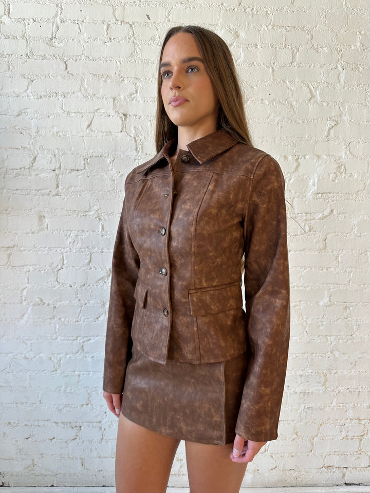 Woman wearing brown leather two piece set with tailored jacket top, and high waisted mini skort bottom.