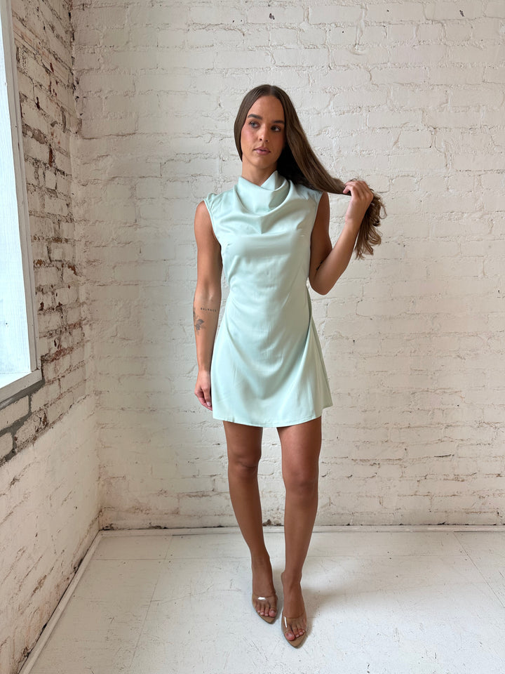 Woman wearing sage green satin mini dress with cap sleeves, and back tie detail.