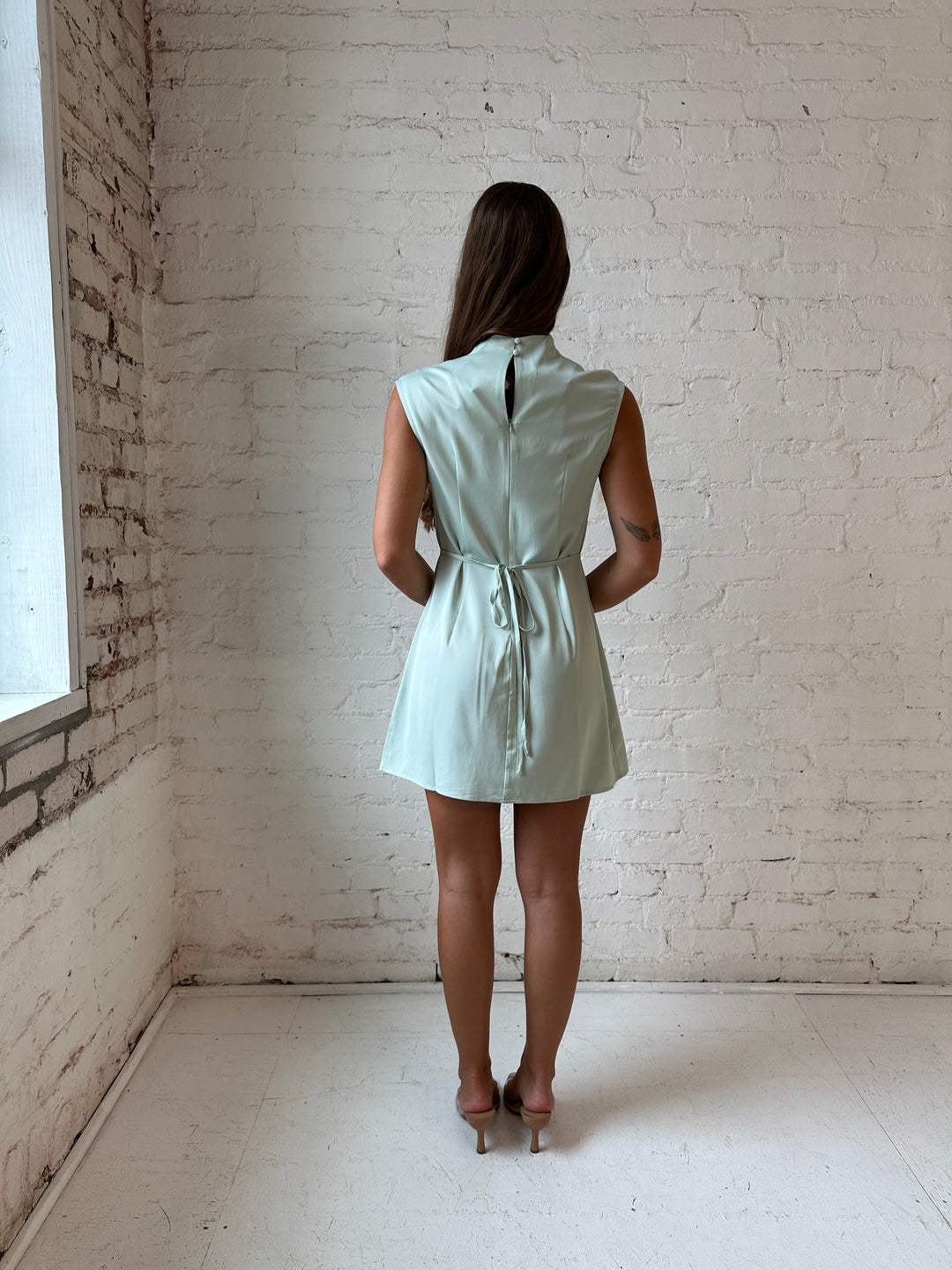 Woman wearing sage green satin mini dress with cap sleeves, and back tie detail.