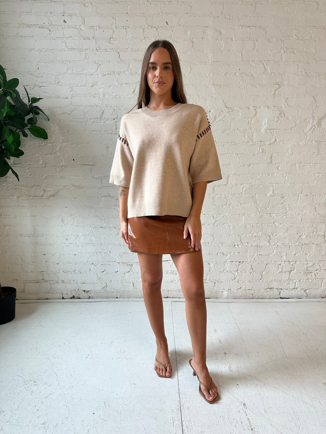 Woman wearing beige colored sweater with contrast stitch sleeve detail and cognac leather mini skirt. 