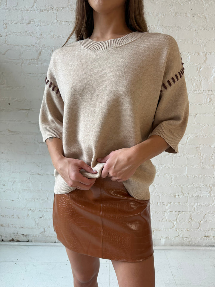 Woman wearing beige colored sweater with contrast stitch sleeve detail and cognac leather mini skirt. 