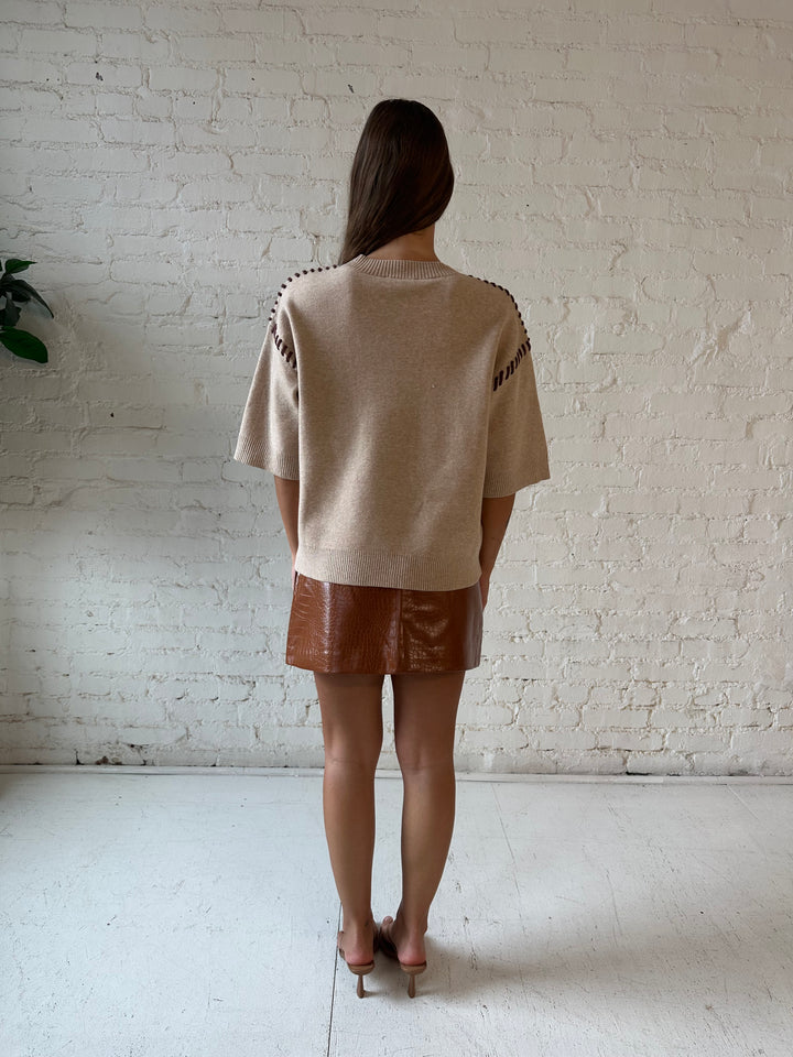 Woman wearing beige colored sweater with contrast stitch sleeve detail and cognac leather mini skirt. 