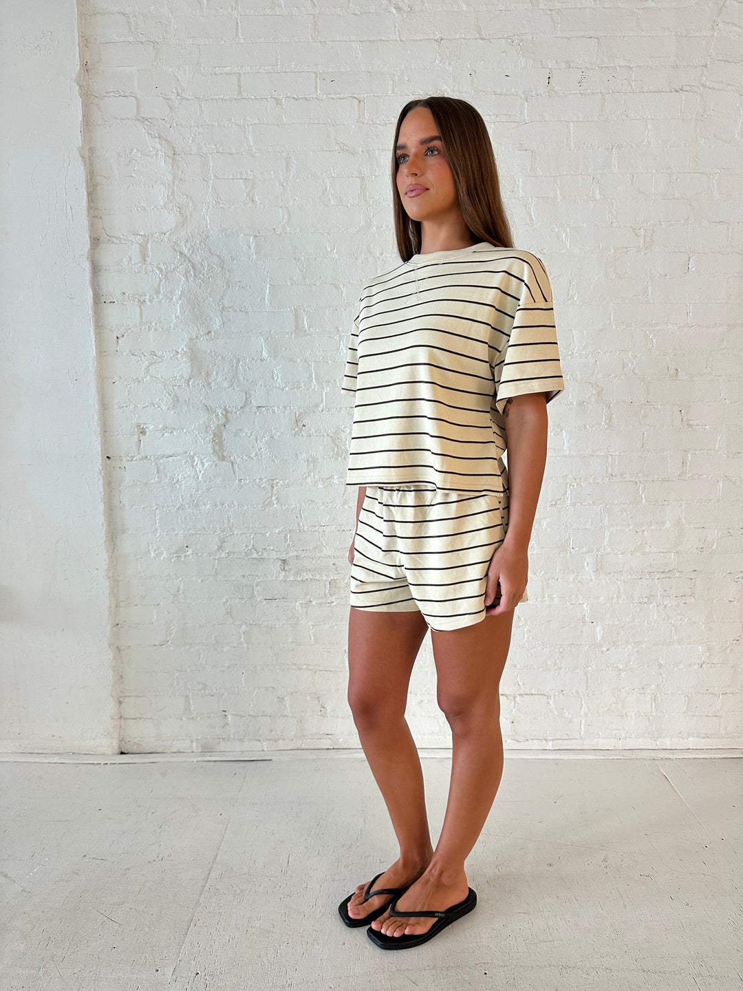 Woman wearing oatmeal and brown striped two piece short lounge set with flip flops. 