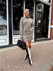 Woman wearing taupe turtle neck sweater dress with belt detail.