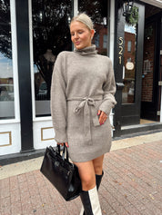 Woman wearing taupe turtle neck sweater dress with belt detail.