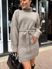 Woman wearing taupe turtle neck sweater dress with belt detail.