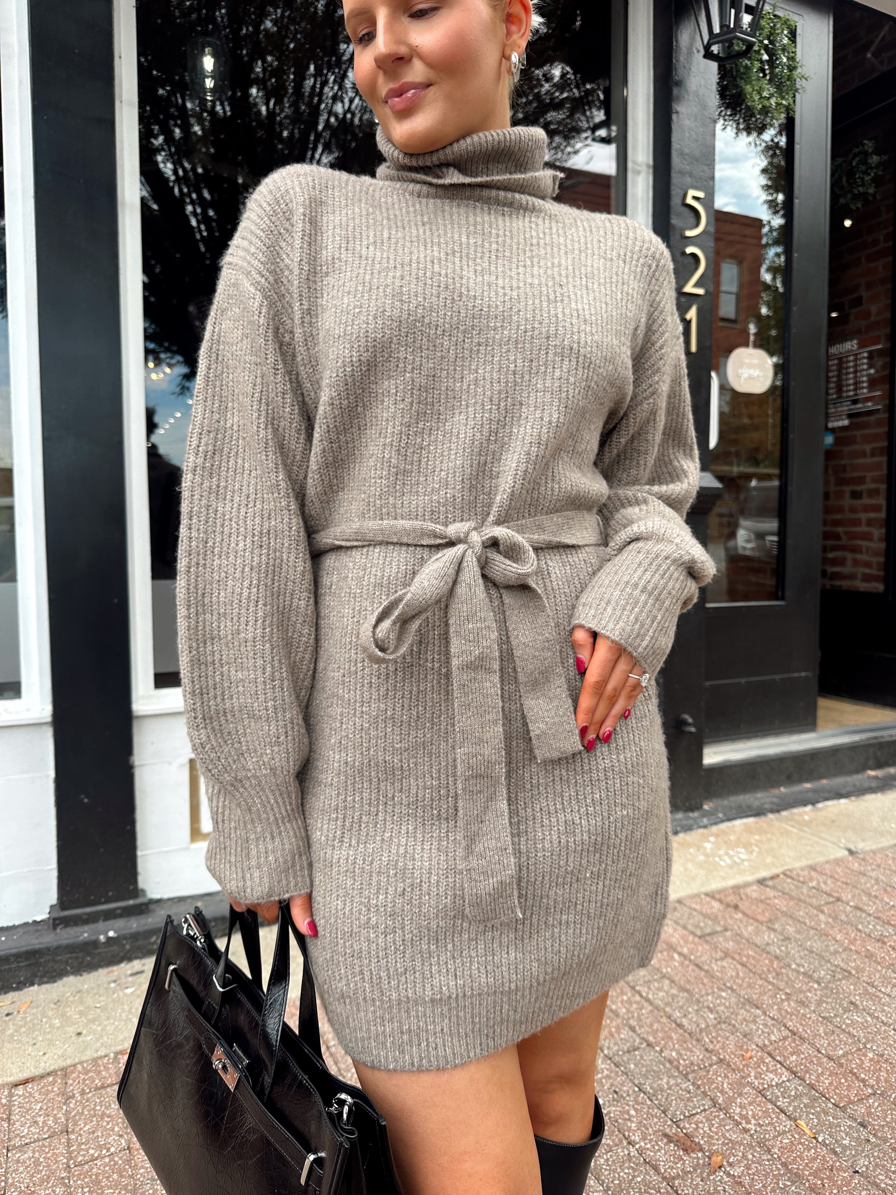 Woman wearing taupe turtle neck sweater dress with belt detail.